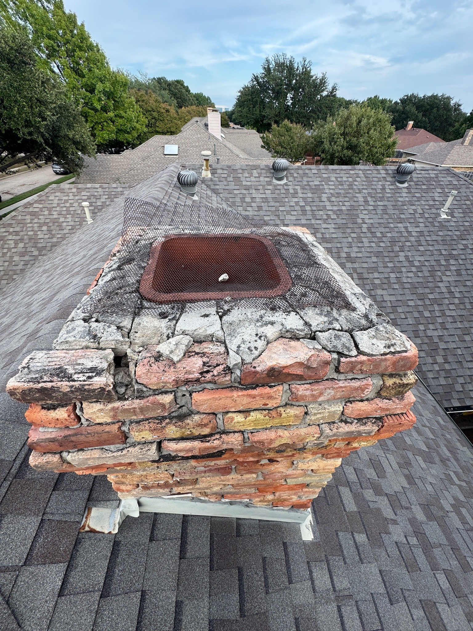 Rooftop view of a brick chimney with a cracked concrete crown, spalling brick, mortar loss and a rusted spark arrestor screen