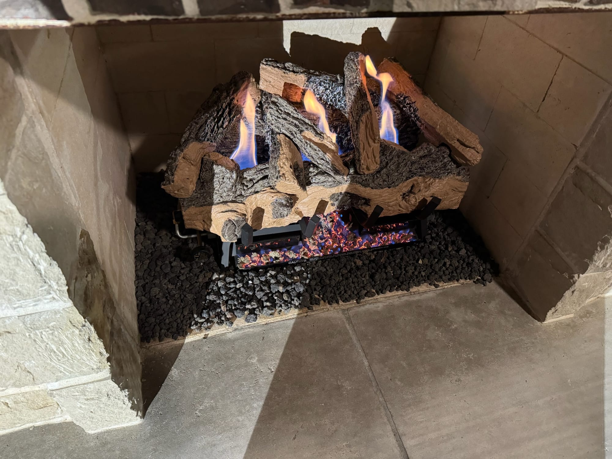 Gas fireplace insert with ceramic logs burning blue and orange flames over black lava rock in a stone firebox