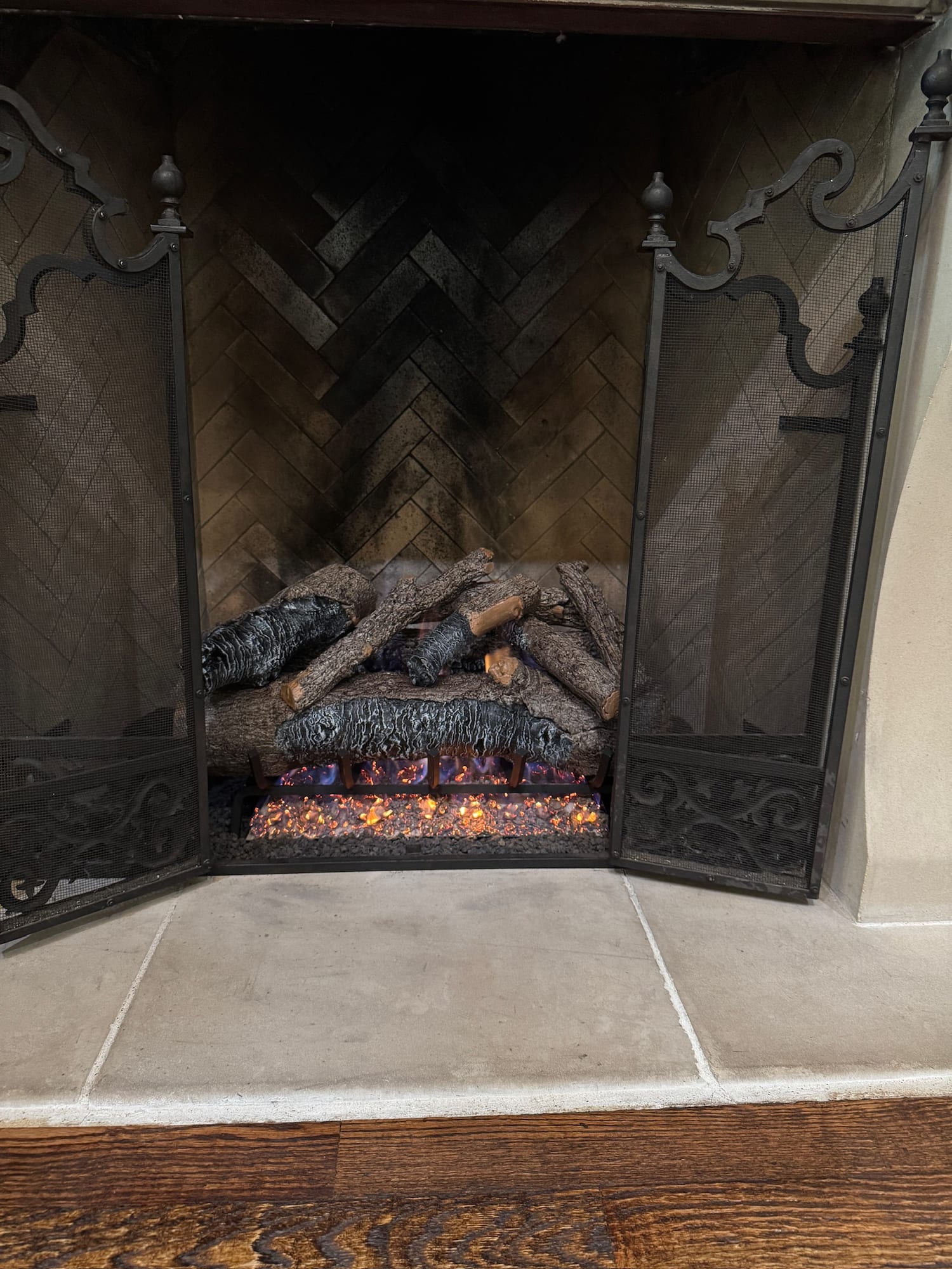 Gas logs burning behind a wrought iron screen in a herringbone brick firebox, soot staining the back wall