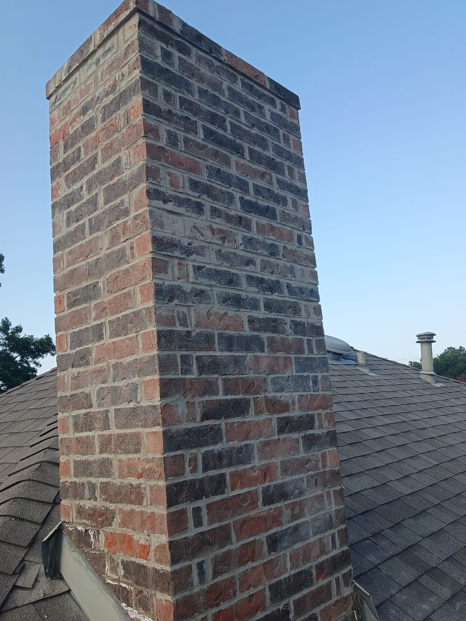 Tall brick chimney with an open, uncapped flue on a shingled roof, with a nearby tree and roof vents under blue sky