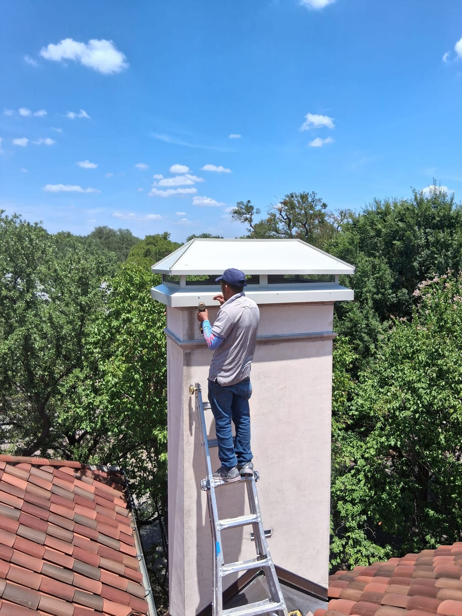 Technician on a ladder examining a large metal cap atop a tall stucco chimney above tiled rooftops