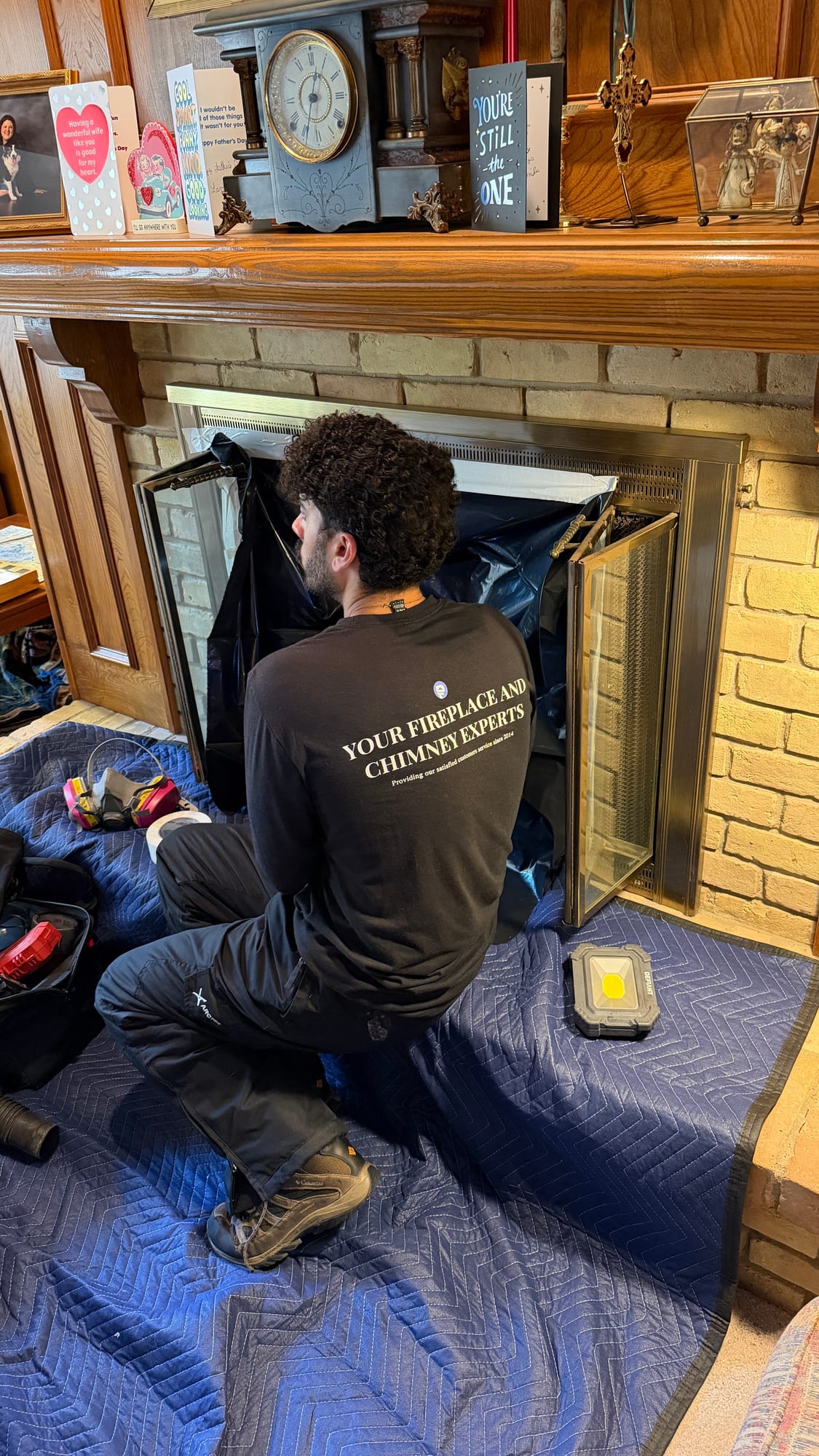 Chimney technician kneeling on a drop cloth servicing an open fireplace insert with a work light under a wooden mantel