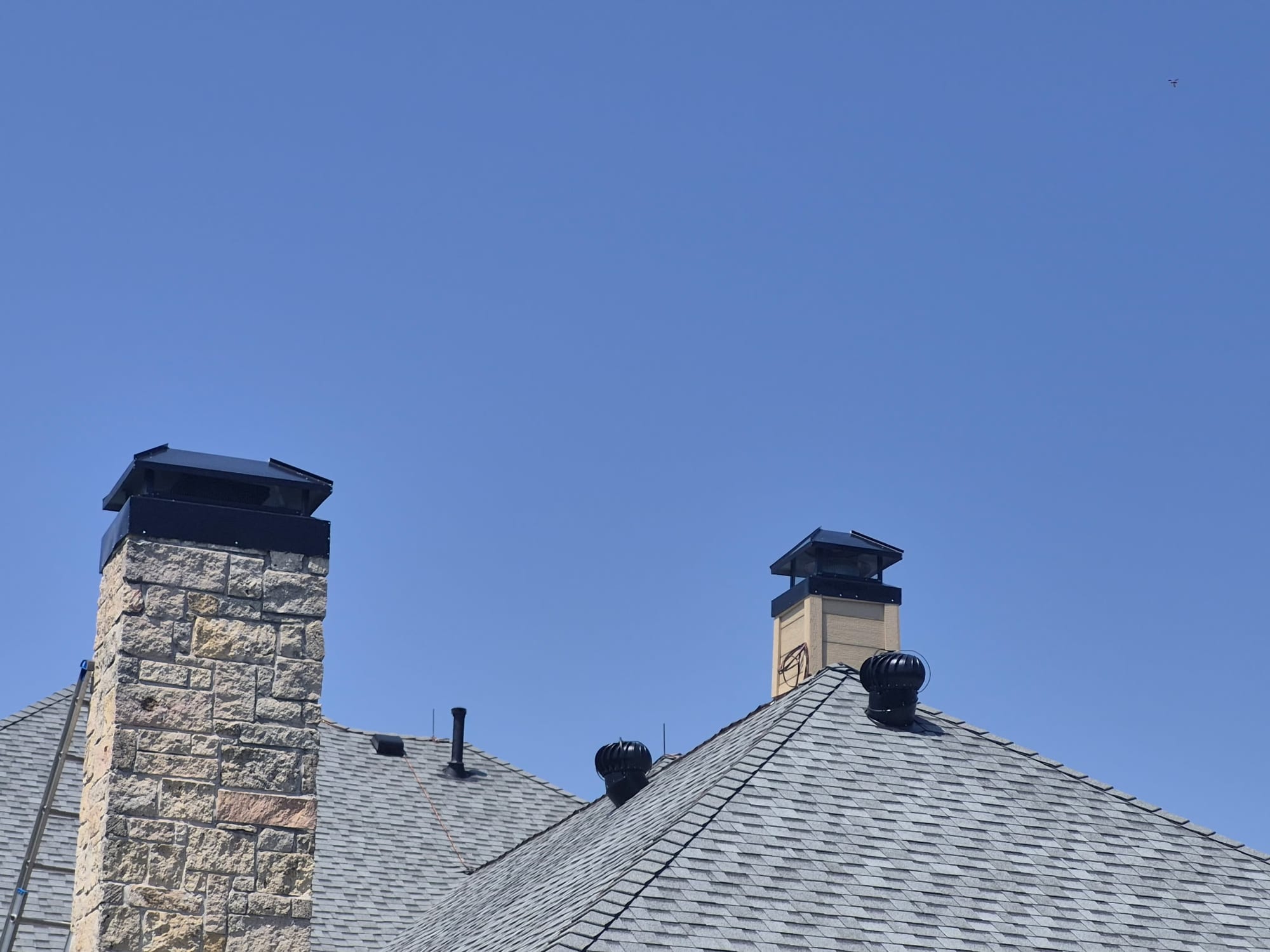 Two rooftop chimneys side by side, one stacked stone and one wood sided chase, each topped with a new black metal cap